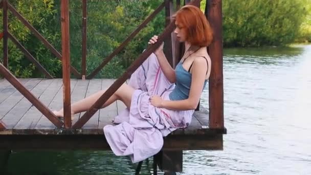 Close-up of beautiful young woman with red hair in blue shirt and long light skirt sitting on a wooden pier and looking at the camera near the river and green trees. Peace and quiet. — Stock Video