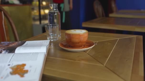 Кофейная кружка с узором на столе в кафе. Искусство. Стол в уютном кафе, на котором стоит кружка кофе и книга. Кофе, кафе и уютный отдых — стоковое видео