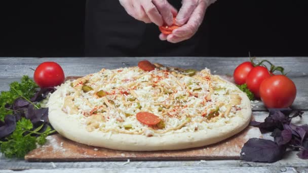 Chefs mãos em luvas de silicone adicionando os pedaços de saveloy em uma pizza que se encontra perto de tomates, salsa e manjericão. Moldura. Deliciosa preparação de pizza — Vídeo de Stock
