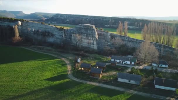 Вид з повітря на зелене поле і маленькі будинки біля схилу гори на блакитне небо. Кадри. Прекрасний літній пейзаж — стокове відео