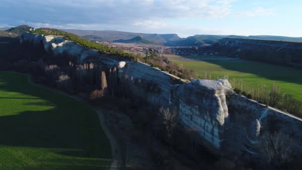 Luftaufnahme von atemberaubenden Natursteinmauer, hohe und lange Klippe mit wachsenden Bäumen und grünen Feldern auf jeder Seite davon. Schuss. wunderschöne Landschaft mit hohen Bergen und einem Dorf im Tal. — Stockvideo