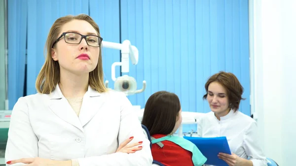 Vrouw in witte medische jas staat voor een camera op tandarts kantoor met een patiënt en een andere arts praten op de achtergrond. De media. Hoofdarts tandheelkunde en haar assistent op het werk. — Stockfoto