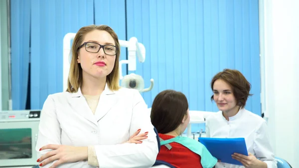 Vrouw in witte medische jas staat voor een camera op tandarts kantoor met een patiënt en een andere arts praten op de achtergrond. De media. Hoofdarts tandheelkunde en haar assistent op het werk. — Stockfoto