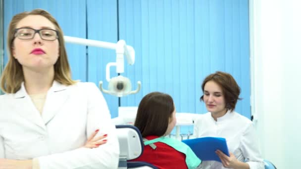 Жінка в білому медичному пальто стоїть перед камерою в стоматологічному кабінеті з пацієнтом та іншим лікарем, який говорить на фоні. Медіа. Головний лікар стоматології та її асистент на роботі . — стокове відео