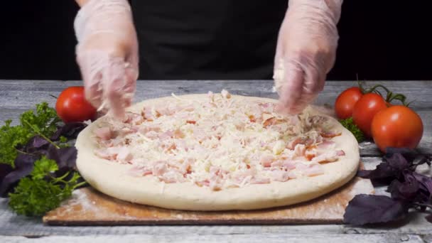 Zavřete ruce v rukavicích, které připravují pizzu ležící na dřevěné desce s čerstvou zeleninou a konceptem pro potravinářství. Rámeček. Kuchař přidává sýr na pizzu na černém pozadí. — Stock video