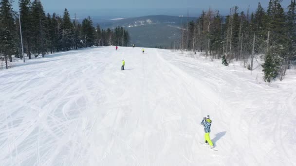 Vista aérea de pessoas em uma pista de esqui começando a descer de uma pista perto de floresta de coníferas no inverno com vista incrível sobre uma montanha coberta por árvores nevadas no fundo. Filmagem. Estância de esqui — Vídeo de Stock