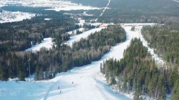 Vista aérea de la pista de esquí y personas haciendo snowboard en una pista de esquí con árboles de coníferas desde ambos lados de la pista. Filmación. Estación de esquí — Vídeos de Stock
