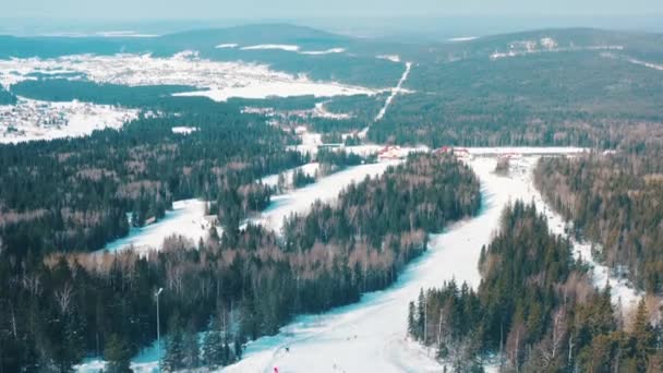 Vista aérea de la pista de esquí y personas haciendo snowboard en una pista de esquí con árboles de coníferas desde ambos lados de la pista. Filmación. Estación de esquí — Vídeos de Stock