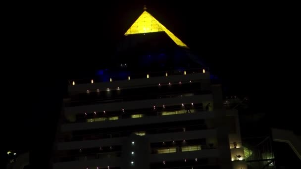 Üstünde sarı piramit olan güzel ve modern bir binanın gece manzarası. Stok. Parlayan piramit çatılı lüks otelin alt görünümü. — Stok video