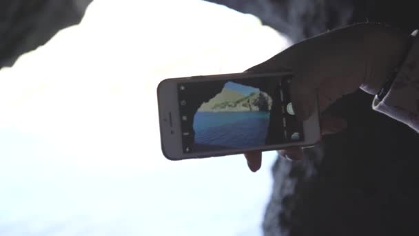 Крупный план туриста фотографирует по телефону пейзаж гор. Искусство. Женская рука с телефоном фотографирует пейзаж моря и скалы с деревьями в пещере — стоковое видео