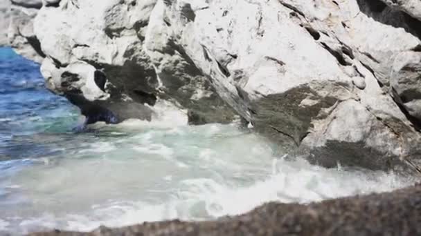 Modré malé vlny se rozdrobí na skály a plážový písek. Článek. Blízko malých pobřežních vln, které se valí na písek a rozbíjí kameny na slunci. Nejlepší dovolená na moři je moře, modré vlny a teplé letní slunce — Stock video