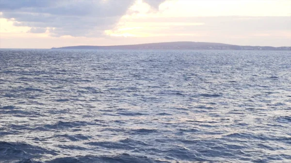 Pintoresca vista de la superficie de agua azul contra la puesta del sol cielo nublado y la orilla del mar en el fondo. Acciones. Impresionante paisaje marino con ondas y olas en un día soleado . — Foto de Stock