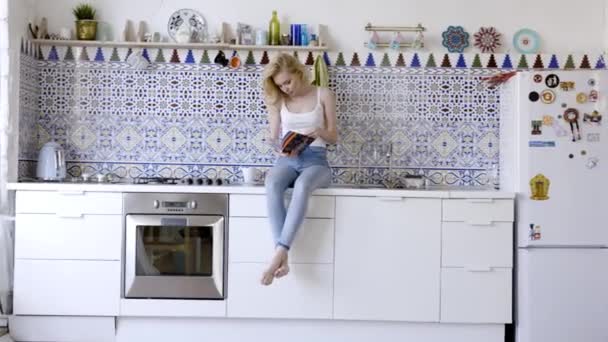 Mirando en la revista por la mañana. Acción. Hermosa joven alegre leyendo revista mientras está sentado en la cocina en casa — Vídeo de stock