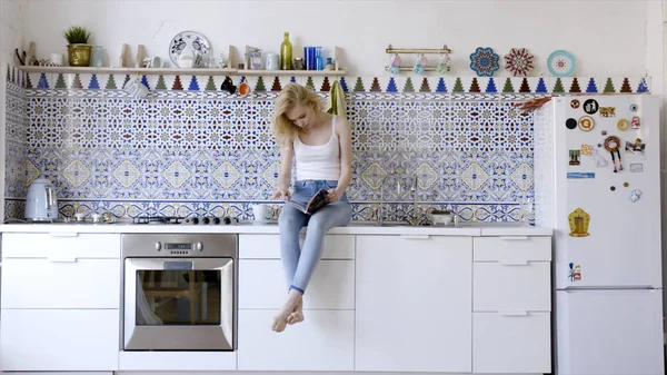 A ver a revista de manhã. Acção. Bela alegre jovem mulher ler revista enquanto sentado na cozinha em casa — Fotografia de Stock