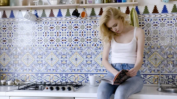 Mirando en la revista por la mañana. Acción. Hermosa joven alegre leyendo revista mientras está sentado en la cocina en casa —  Fotos de Stock