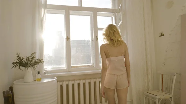 Fiatal és gyönyörű szőke nő pózol az ablak mellett. Felvétel. Napfény az ablakból. Szexi szőke a nappaliban reggel — Stock Fotó