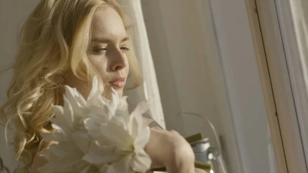 Mujer rubia joven con flores en las manos al sol. Acción. Joven hermosa mujer en la luz del sol en casa con flores — Foto de Stock