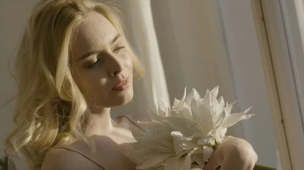 Young blonde woman with flowers in her hands in the sun. Action. Young beautiful woman in sunlight at home with flowers