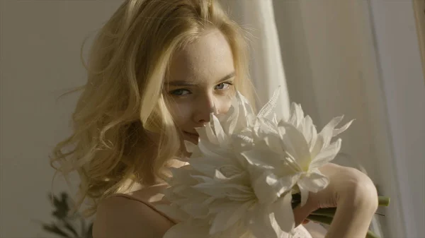 Mujer rubia joven con flores en las manos al sol. Acción. Joven hermosa mujer en la luz del sol en casa con flores — Foto de Stock