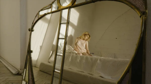 Krásná blondýnka v noční košili na bílé posteli ohýbá nohy. Akce. Odraz v zrcadle — Stock fotografie