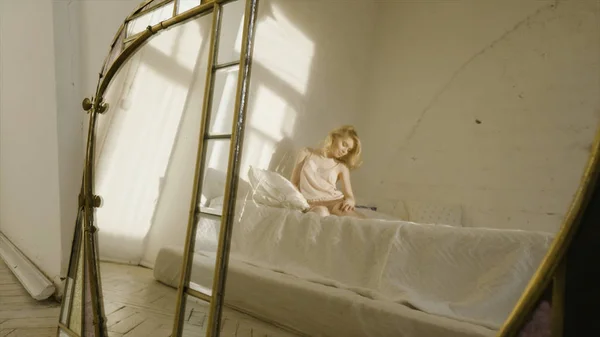 Krásná blondýnka v noční košili na bílé posteli ohýbá nohy. Akce. Odraz v zrcadle — Stock fotografie
