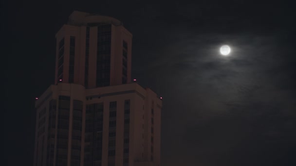 Night cityscape with a high building on dark sky background. Stock footage. Full moon on dark cloudy sky. — Stock Video