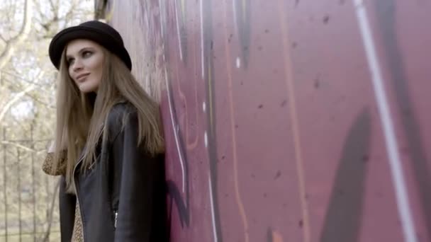 黒い帽子をかぶったセクシーなモデルの女性は、壁の近くに立ってポーズをとる。アクション。壁の近くに立っている帽子をかぶったスタイリッシュな若い女性 — ストック動画