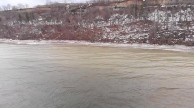 Geniş nehrin havadan görünümü ve tepede yetişen ağaçlarla karlı kıyı, kışın doğa. Stok. Soğuk sarı nehir ve ormanlık dağ ile kış manzara.