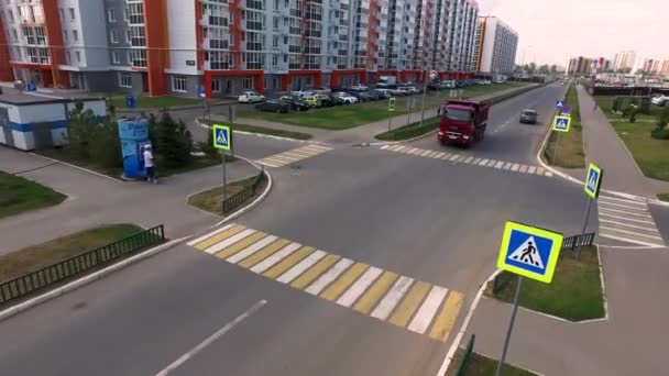 Aérea para el gran camión rojo que se mueve en la calle de un distrito moderno de la ciudad con casas de varios pisos en el fondo. Escena. Conducción de camiones en la encrucijada con paso peatonal . — Vídeos de Stock