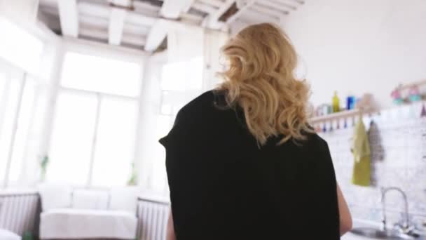 Close-up of beautiful blond woman in white t-shirt, jeans and black jumper going towards the window in her appartments. Action. Relaxing at home — Stock Video