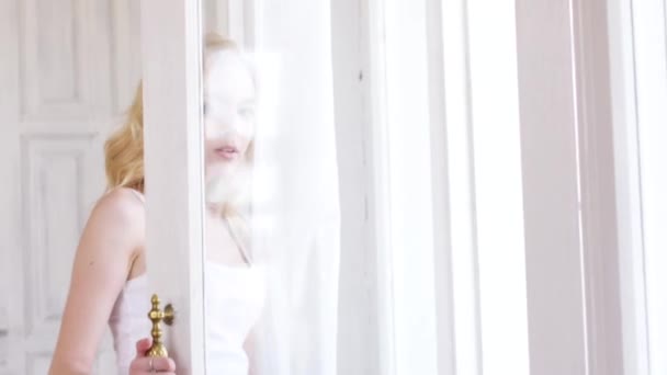 Gros plan de la belle jeune femme aux yeux bleus en t-shirt blanc debout près de la fenêtre et regardant la caméra. L'action. Détente à la maison — Video
