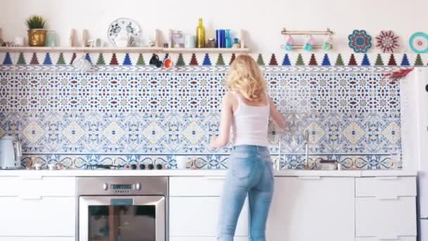 白いTシャツと青いジーンズでブロンドの髪を持つ美しい若い女性の背中から、キッチンでテーブルを磨きます。アクション。家事 — ストック動画