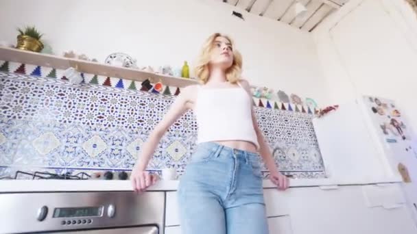 Beyaz tişörtlü sarı saçlı ve mutfak alanında duran ve kameraya bakan mavi kot pantolonlu güzel genç kadının yakından görünümü. Eylem. Evde rahatlatıcı — Stok video