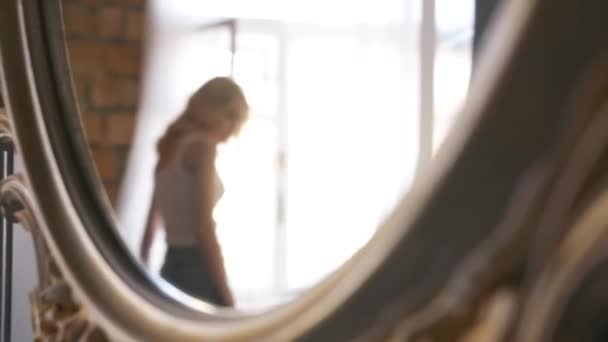 白いTシャツと青いジーンズを着た若いブロンドの女性が窓の近くを歩いている鏡の鏡の鏡の鏡の鏡の鏡の鏡の鏡の鏡の反射。アクション。自宅でリラックス — ストック動画