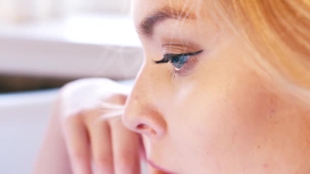 Close-up de mulher loira bonita com olhos azuis profundos e maquiagem perfeita sentado no banheiro e pensando em algo. Acção. Relaxar em casa — Vídeo de Stock