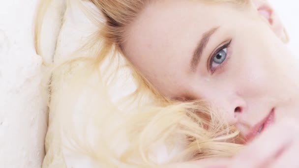 Close-up of beautiful young womans face with perfect makeup, blonde hair and blue-and-grey eyes lying on soft and white pillow. Action. Women images concept — Stock Video