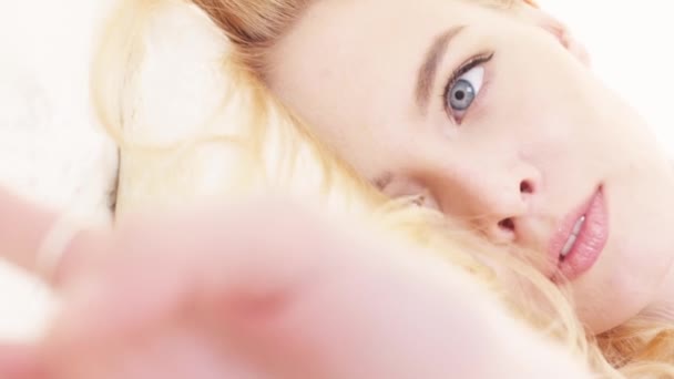 Primer plano de la hermosa cara de las mujeres jóvenes con maquillaje perfecto, cabello rubio y ojos azules y grises acostados sobre una almohada suave y blanca. Acción. Mujeres imágenes concepto — Vídeos de Stock