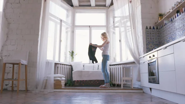 Nahaufnahme einer schönen blonden Frau in weißem T-Shirt und Jeans, die am Fenster steht und in ihren Appartements einen schwarzen Pullover anzieht. Action entspannt zu Hause — Stockfoto