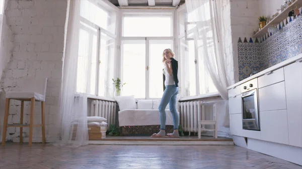 Nahaufnahme einer schönen blonden Frau in weißem T-Shirt und Jeans, die am Fenster steht und in ihren Appartements einen schwarzen Pullover anzieht. Action entspannt zu Hause — Stockfoto