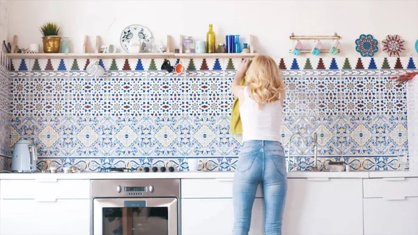 Blick von der Rückseite der schönen jungen Frau mit blonden Haaren in weißem T-Shirt und blauer Jeans, die den Tisch in der Küche poliert. Hausarbeit — Stockfoto