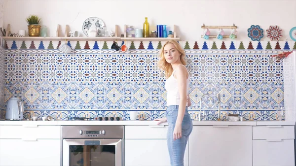 Zblízka krásná mladá žena se světlými vlasy v bílých a modrých džínách, které dělají čaj nebo kávu v kuchyni. Akce. Relaxace doma — Stock fotografie