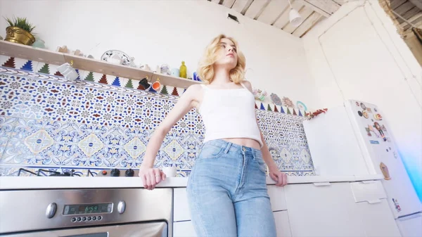 Beyaz tişörtlü sarı saçlı ve mutfak alanında duran ve kameraya bakan mavi kot pantolonlu güzel genç kadının yakından görünümü. Eylem. Evde rahatlatıcı — Stok fotoğraf