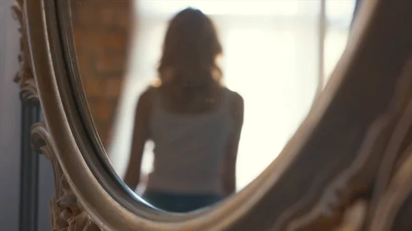 Primer plano del espejo reflejo de la joven rubia en camiseta blanca y jeans azules caminando cerca de una ventana en sus departamentos. Acción. Relajarse en casa — Foto de Stock