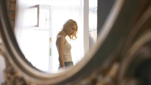 Close-up de espelho reflexo de mulher loira jovem em t-shirt branca e jeans azul andando perto de uma janela em seus compartimentos. Acção. Relaxar em casa — Fotografia de Stock