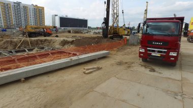 Ağır kırmızı kamyon arka planda diğer makinelerle çok katlı binanın inşaat yerine geliyor. Sahne. Bir kamyon, kamyon ve yüksek katlı vinç ile modern bina inşaatı.