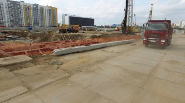 Ağır kırmızı kamyon arka planda diğer makinelerle çok katlı binanın inşaat yerine geliyor. Sahne. Bir kamyon, kamyon ve yüksek katlı vinç ile modern bina inşaatı.