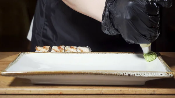 Närbild för händer i svart matlagning handskar klämma grön Wasabi sås på en tallrik förberedda rullar. Ram. Japanska skålen förberedelse, foodporn Concept. — Stockfoto