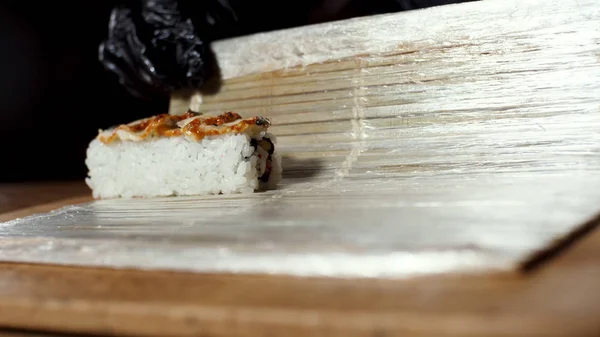 Prohlédněte si šéfkuchaře černými kuchyňskými rukavicemi a Suši makisu, japonský potravinový přípravek. Rámeček. K ruce si zavání sushi s tradiční bambusovou rohoží. — Stock fotografie