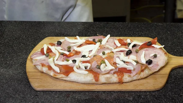 O chef em luvas de cozinha preta fazendo pizza deitada em uma tábua de madeira, conceito de comida deliciosa. Moldura. Fechar para as mãos colocando azeitonas e mozarella na pizza . — Fotografia de Stock