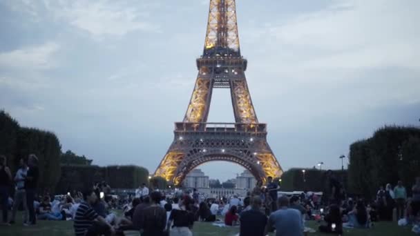 A csillogó Eiffel-torony turistái este. Akció. Sok turista ül a gyepen, és élvezze a gyönyörű kilátás a csillogó Eiffel torony este nyáron — Stock videók